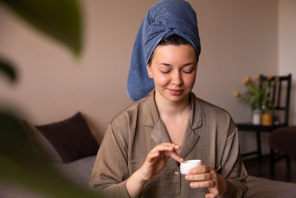 Woman with a hair towel scooping face moisturizer from its jar