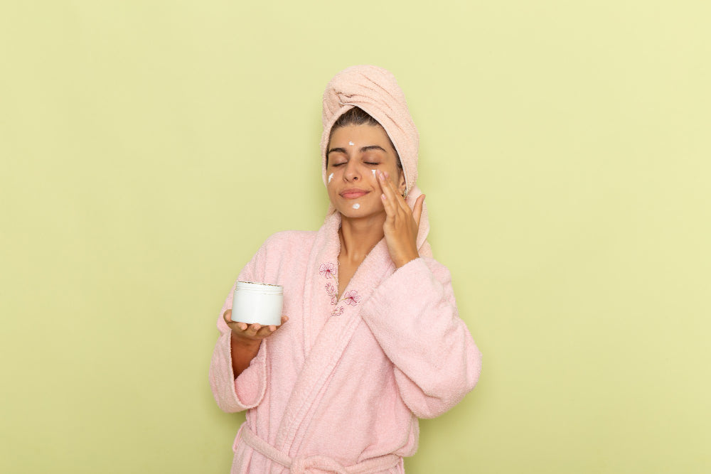 Woman in a pink robe applying face moisturizer from a jar