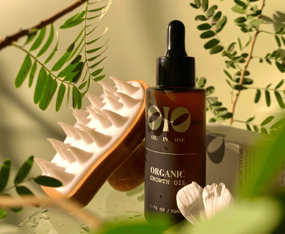 Oil In One Organic Growth Oil bottle with a scalp massager brush among leaves